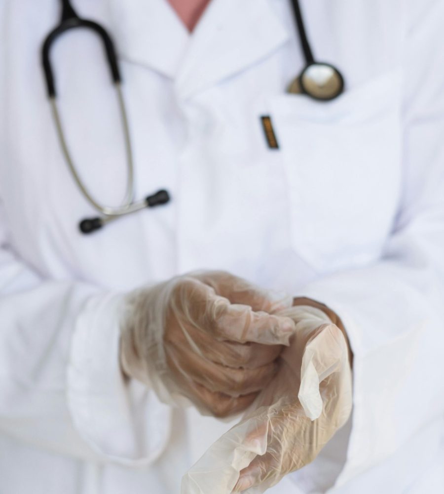 Faceless ethnic medical worker in lab coat and stethoscope taking of transparent gloves after approaching patients for examination in modern hospital